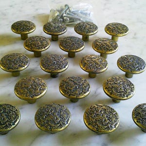 May include: A set of 16 antique-style cabinet knobs. Each knob has a textured, dark bronze finish with a gold-colored rim. The knobs are arranged on a white marble surface, with a small bag of screws visible in the background.
