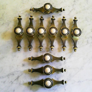 May include: A collection of ten drawer pulls featuring an antique brass finish with a central circular white ceramic inlay. The hardware consists of six vertical pulls and four horizontal pulls, each with a decorative metal frame and a smooth finish.