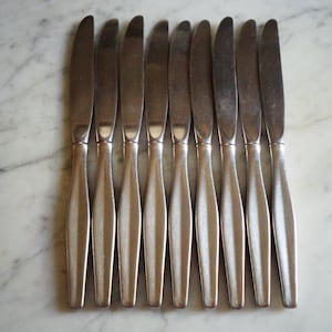 May include: A set of nine silver-toned stainless steel dinner knives arranged in a parallel row. Each knife features a smooth, curved blade and a handle with a central vertical ridge, showcasing a polished metallic finish throughout the design.