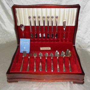 May include: A wooden box with a red velvet interior containing a set of Community silver cutlery. The box is open and the cutlery is arranged in rows. The cutlery includes forks, knives, and spoons.
