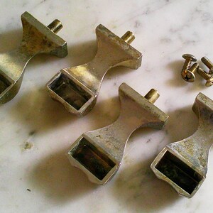 Op de afbeelding: Vier zilverkleurige meubelknoppen van metaal met een taps toelopende, zandloperachtige vorm en een rechthoekige basis. Elke knop heeft een metalen schroefdraadpen die aan de bovenkant uitsteekt. Drie kleine, bijpassende zilverkleurige metalen schroeven zijn inbegrepen.