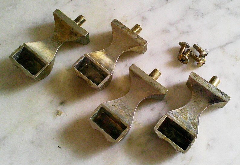 Op de afbeelding: Vier zilverkleurige meubelknoppen van metaal met een taps toelopende, zandloperachtige vorm en een rechthoekige basis. Elke knop heeft een metalen schroefdraadpen die aan de bovenkant uitsteekt. Drie kleine, bijpassende zilverkleurige metalen schroeven zijn inbegrepen.