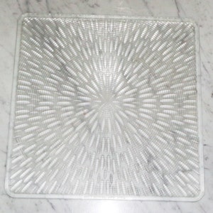 Puede incluir: Una alfombra de ducha de plástico transparente y cuadrada con una superficie texturizada. La textura es un patrón repetitivo de pequeñas formas ovaladas que irradian hacia afuera desde el centro de la alfombra.