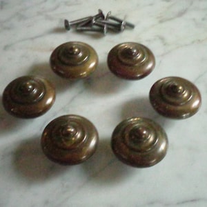 Peut inclure: Six boutons de cabinet en laiton antique avec un motif central surélevé. Les boutons sont ronds et ont une forme légèrement bombée.