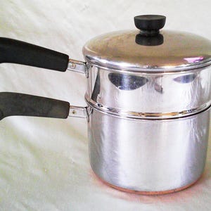 May include: A stainless steel double boiler with a polished finish, featuring two stacked cylindrical pots, a matching lid with a black knob, and two black handles, each equipped with a small metal hanging loop at the end of the handle.