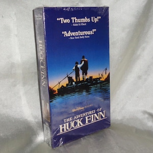 Può includere: Una custodia per videocassetta VHS per il film "Le avventure di Huckleberry Finn". La custodia è blu e presenta una scena del film con due ragazzi su una barca su un fiume. La custodia include anche citazioni di critici, tra cui "Due pollici in su!" e "Avventuroso!"