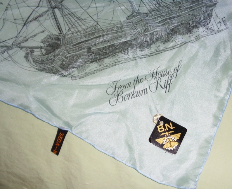 May include: A light blue silk scarf featuring a detailed black and white illustration of a sailing ship. The scarf has the text "From the House of Borkum Riff" and a tag that reads "B.N."