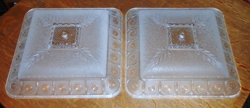 May include: Two frosted glass ceiling light fixtures with a square shape and a decorative pattern. The fixtures have a frosted glass center with a square design and a decorative border.
