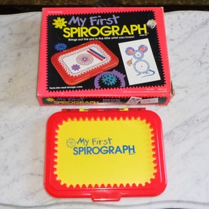May include: A red and yellow plastic case with the words "My First Spirograph" on the front. The case is closed and has a scalloped edge. The case is a toy for drawing geometric shapes.