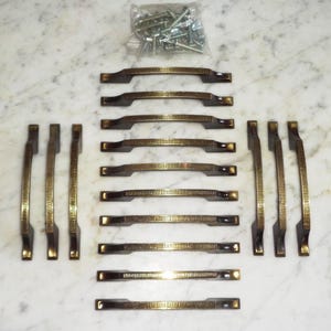 May include: A collection of bronze-toned cabinet pulls and matching vertical handles, arranged on a marble surface. The pulls have a rectangular design with a textured center, and the handles have a similar textured pattern. A small bag of screws is also included.