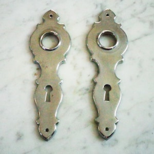 May include: Two silver door plates with ornate designs and keyholes. The plates are rectangular with a rounded top and bottom. Each plate has a circular cutout for a door knob and a keyhole in the center.