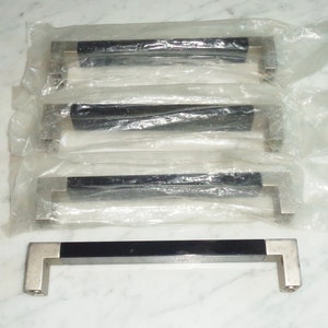 Puede incluir: Cuatro tiradores de armario negros con detalles plateados en envases de plástico individuales. Los tiradores son rectangulares con bordes redondeados y una ligera curva en el centro.