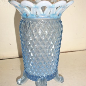 Puede incluir: Un jarrón de vidrio azul claro con un patrón de diamante y un borde de encaje blanco. El jarrón tiene tres patas y mide aproximadamente 20 cm de alto.