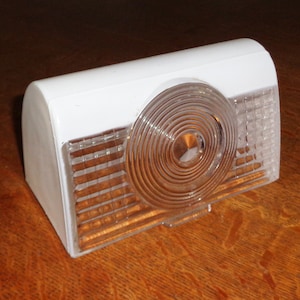 May include: White plastic automotive taillight with a clear lens and a circular reflector. The lens has a grid pattern and the reflector has concentric circles.