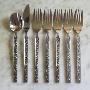 May include: A set of vintage silverware, including a spoon, knife, and six forks. The stainless steel utensils have ornate, floral-patterned handles. The silverware is arranged on a light-colored, marble-like surface.