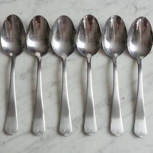 May include: Six silver teaspoons with ornate handles. The spoons are arranged in a row on a white marble surface.