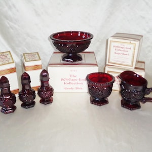 Op de afbeelding: Een set rode glazen Cape Cod-collectieartikelen, waaronder een snoepschotel, een suikerpot, een roomkan en zout- en peperstrooiers. De artikelen bevinden zich allemaal in hun originele dozen.