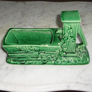 May include: A green ceramic planter shaped like a well with a pump handle. The planter has a rectangular base and a raised well structure with a pump handle on the side. The planter is decorated with a rustic wood grain pattern and vines.