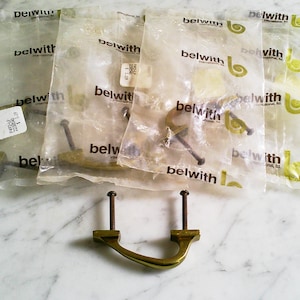 May include: Several brass-toned cabinet pulls, some still in their original plastic packaging. The packaging has the brand name "belwith" printed on it. One pull is displayed outside the packaging, showing its curved handle and mounting screws.