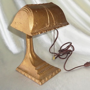 Puede incluir: Una lámpara de escritorio de metal de color dorado con una pantalla que tiene un diseño decorativo. La lámpara tiene una cadena de tracción y un cable con un enchufe.