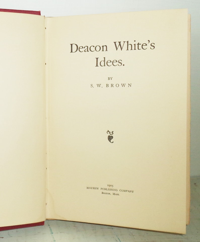 May include: Open book with the title "Deacon White's Idees" by S.W. Brown. The text is in a serif font, printed in brown ink on aged, cream-colored paper. The book was published in 1905 by Mayhew Publishing Company, Boston, Mass.