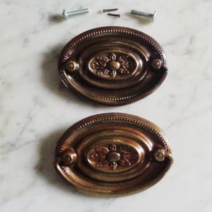May include: Two antique bronze oval drawer pulls with a floral design in the center. The pulls have a beaded edge and are accompanied by screws for installation. The hardware is set against a white marble surface.