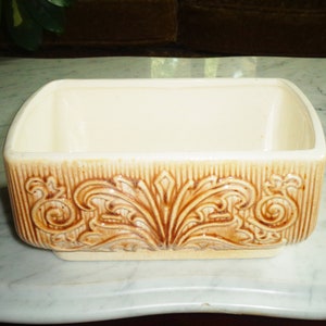 May include: A rectangular ceramic planter with a white glaze and brown floral design. The planter has a raised base and a smooth interior.