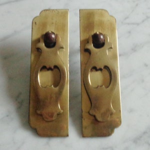 May include: Two antique brass drawer pulls with a decorative, ornate design. Each pull features a rounded top with a small, round knob and a cutout in the shape of a vase.