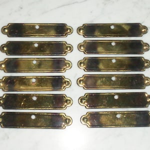 Peut inclure: Dix tirettes de tiroir en laiton antique de forme rectangulaire arrondie avec un seul trou au centre. Les tirettes ont une finition légèrement ternie.
