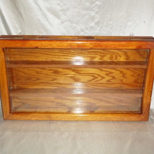 May include: A wooden display case with three shelves and a glass front. The case is made of light brown wood with a natural grain pattern. It has a hinged top and two small metal hooks on the top for hanging.