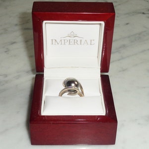 Pode incluir: Um anel de ouro com uma p&eacute;rola cinza escura e pequenos diamantes engastados na banda. O anel est&aacute; em uma caixa de anel de veludo vermelho com a palavra "IMPERIAL" impressa no interior da tampa.