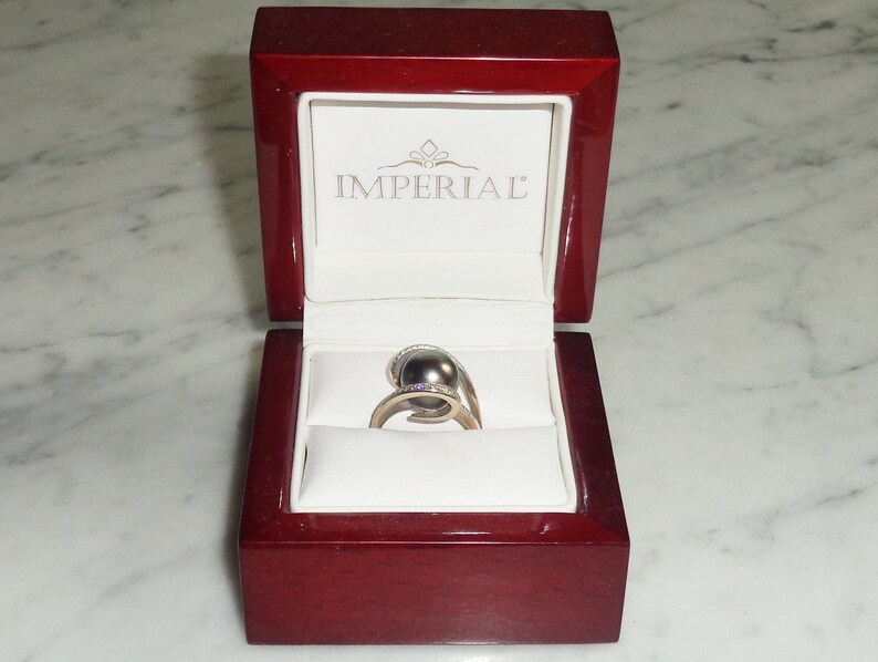 Pode incluir: Um anel de ouro com uma p&eacute;rola cinza escura e pequenos diamantes engastados na banda. O anel est&aacute; em uma caixa de anel de veludo vermelho com a palavra "IMPERIAL" impressa no interior da tampa.