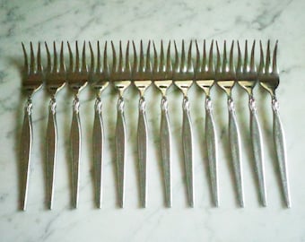 Op de afbeelding: Een verzameling van twaalf zilveren vorken, gerangschikt in een rij. Elke vork heeft een slanke steel met een decoratief bloemmotief aan de basis van de tanden. De vorken worden gepresenteerd op een lichtgekleurd, marmerachtig oppervlak.