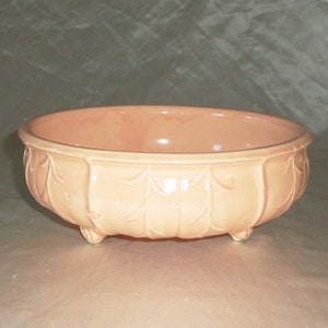 Puede incluir: Un macetero de cerámica de color melocotón pálido con un borde festoneado y un diseño de hojas. El macetero tiene tres pequeños pies y es perfecto para exhibir plantas.