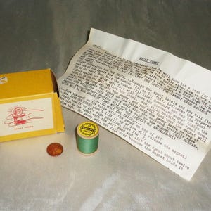 May include: A yellow box with a red and white illustration of a hand holding a spool with a penny on top. The box is labeled "Macky Punkt". There is a spool of green thread with a yellow label that reads "Penny Size" and a copper penny next to the box. A piece of paper with instructions for a magic trick is visible in the background.