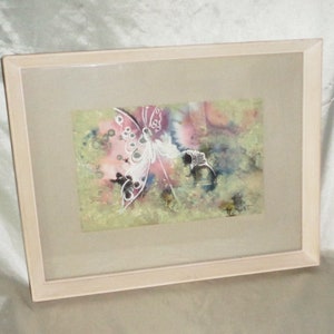 May include: A framed watercolor painting of a white butterfly with black dots on its wings. The butterfly is flying over a pink and green background with a signature in the bottom right corner.