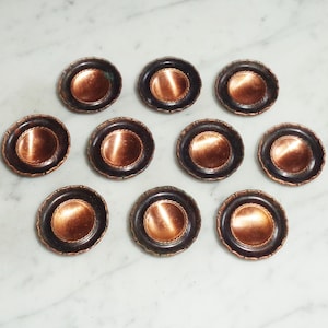Könnte beinhalten: Zehn kleine, runde Untersetzer in Kupferfarbe mit schwarzen Rändern. Die Untersetzer sind in einem Kreis auf einer weißen Marmorfläche angeordnet.