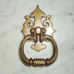 Puede incluir: Un llamador de puerta de metal dorado con un diseño decorativo y ornamentado. El llamador tiene un asa en forma de anillo y una parte superior decorativa.