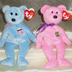 May include: Two plush Beanie Baby bears, one light blue and one pink, each with a heart-shaped tag. The bears have embroidered features and an Easter egg design on their chests. Each bear has a ribbon around its neck.
