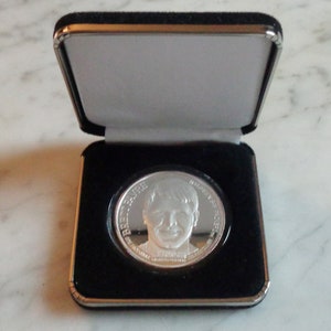 May include: A silver coin featuring a portrait of a person, displayed in an open black presentation box with a gray interior. The coin has text around the portrait. The box has a silver trim.