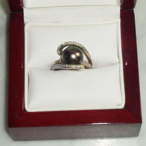 Pode incluir: Um anel de ouro branco com uma p&eacute;rola preta e um design de espiral cravejado de diamantes. O anel &eacute; apresentado numa caixa de anel de veludo vermelho.