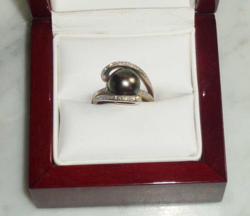 Pode incluir: Um anel de ouro branco com uma p&eacute;rola preta e um design de espiral cravejado de diamantes. O anel &eacute; apresentado numa caixa de anel de veludo vermelho.