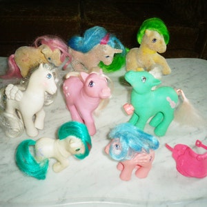 May include: A collection of eight vintage My Little Pony toys, including a pink pony with a blue mane, a white pony with a pink mane, a green pony with a yellow mane, a brown pony with a green mane, a pink pony with a purple mane, a blue pony with a pink mane, a white pony with a blue mane, and a pink saddle.