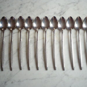 May include: A set of twelve silver teaspoons with a textured handle. The spoons are arranged in a row on a white marble surface.