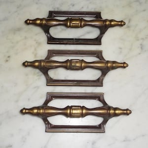 May include: Three antique brass drawer pulls with ornate designs. Each pull has a rectangular plate with a raised center section and a curved handle. The pulls are in good condition with some signs of age.