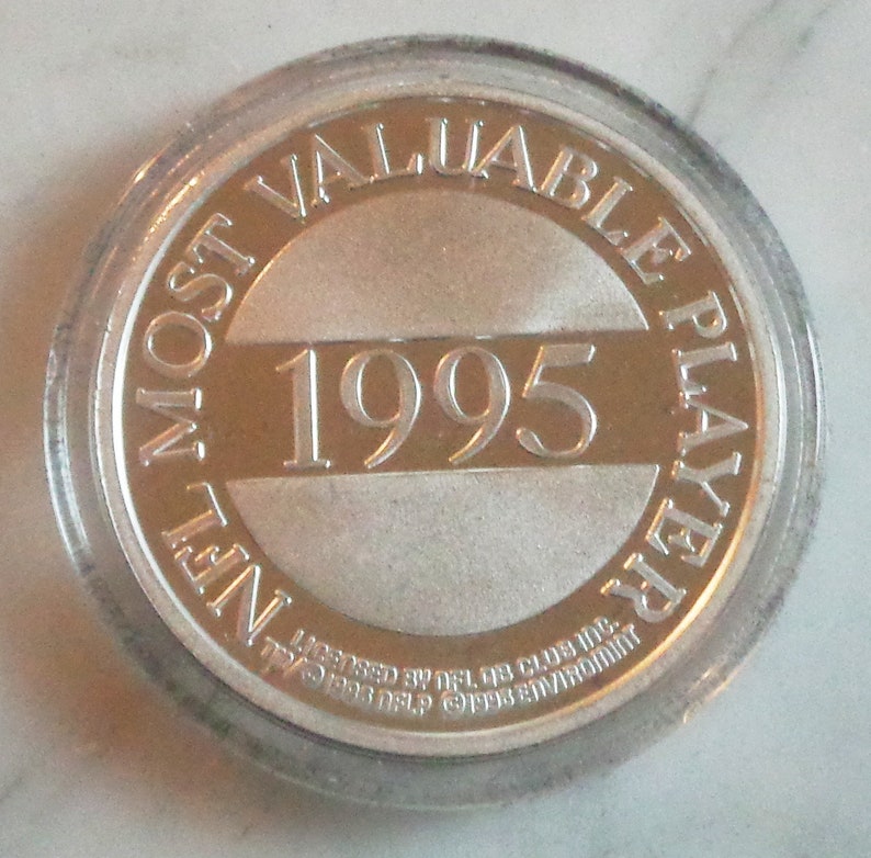 May include: A silver coin encased in a clear plastic holder. The coin features the text "MOST VALUABLE PLAYER" and "1995" in the center. The coin also has the text "NFL" and other text around the edge.
