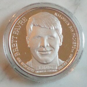 May include: A silver coin featuring a portrait of Brett Favre, with the text "BRETT FAVRE" and "NO. 4 GREEN BAY PACKERS". The coin is marked "ONE TROY OUNCE" and "999 FINE SILVER", with "1806 LIMITED EDITION".