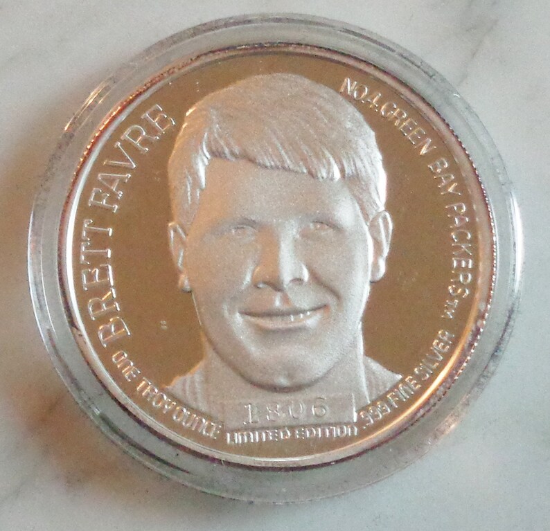 May include: A silver coin featuring a portrait of Brett Favre, with the text "BRETT FAVRE" and "NO. 4 GREEN BAY PACKERS". The coin is marked "ONE TROY OUNCE" and "999 FINE SILVER", with "1806 LIMITED EDITION".