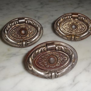 以下が含まれることがあります： 装飾的なデザインが施された、アンティークな楕円形のキャビネットプルが3つ。プルは銀色の仕上げの金属製で、茶色の緑青があります。
