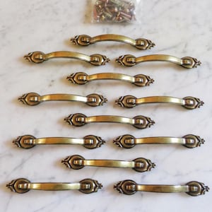 May include: Ten antique brass cabinet pulls with a decorative design. The pulls are approximately 7.5 cm long and have a curved shape.
